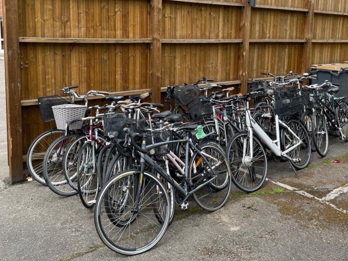 Approximately 25 bicycles stacked against a wooden wall in an unspecified inner courtyard in Copenhagen. The bibycles are ready to be refurbished and used again